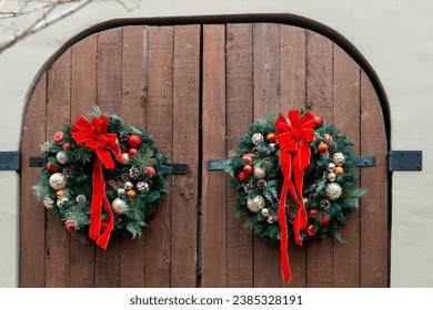 Two brown wooden arch shaped exterior doors on a concrete wall. The door has two black metal hinges. Two Christmas wreaths are hanging on the door. The wreaths have red ribbons, cones and gold balls.  - Powered by Shutterstock