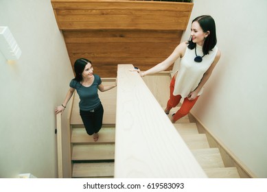 Two Brunette Woman Going Up And Down Upstairs.  On The Stairs And Looking Each Other. Smiley Face. Couple Of Girls Against Wooden Wall With Green Grass. Lesbian Couple Inside Home