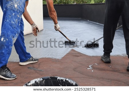 Two builders construction workers with brushes applying waterproofing layer coating on a building roof 
