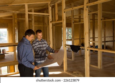 Two builders standing with an open blueprint discussing the interior of a half completed timber frame house - Powered by Shutterstock