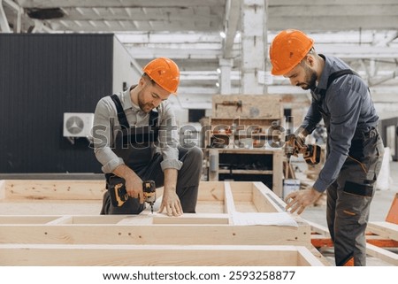 Two builders using cordless screwdrivers assembling prefabricated wooden panels for modular building in large carpentry workshop