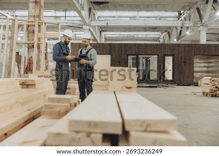 Two builders using a tablet and inspecting wooden planks in a factory for the production of modular buildings