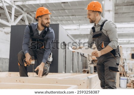 Two builders working on modular building project, using electric screwdriver and checking blueprint in large carpentry workshop