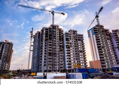 Two Building Cranes At The Background Of A Multi-storey Building Under Construction With Some Small Workers Building In Front Of Them Against Sunset Sky. New Houses Will Wait For Families