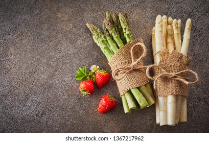 Two bundles of fresh white and green asparagus spears lying on a textured grey background with copy space and three ripe strawberries