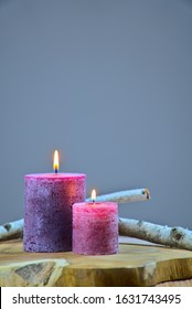 Two Burning Colorful Candles On Gray Background, Wooden Table And Pieces Of Birchwood In The Background.