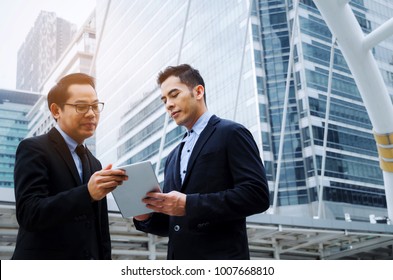 Two Business Asian Smart Man In Suit Talking And Reading Information About Finance News In Mobile Tablet Together Standing In Modern City, Network Technology, Internet, Successful, Teamwork Concept