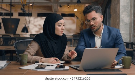 Two business colleagues co-workers talk discuss corporate work at office cafe multiracial male female employees Indian man muslim woman in hijab diverse businesswoman businessman write tasks laptop - Powered by Shutterstock