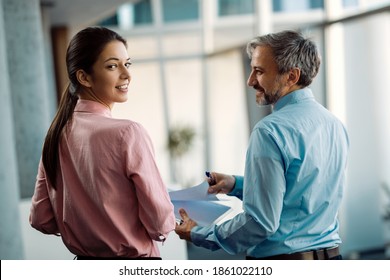 Two Business Colleagues Going Through Paperwork In A Hallway. Focus Is On Young Businesswoman Turning Towards The Camera. 