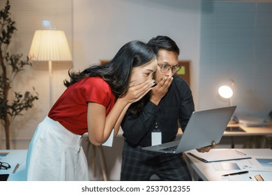 Two business colleagues looking shocked while working late in the office and reacting to something on a laptop - Powered by Shutterstock