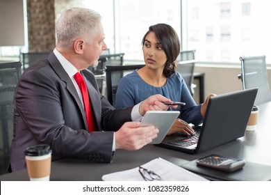 Two Business Colleagues Working Together In Meeting Space. Mentor Instructing Newcomer. Young Indian Woman With Laptop And Mid Adult Man With Tablet Discussing New Project.