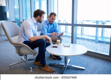 Two Business Entrepreneurs Sitting At A Coffee Table Together In A Bright Modern Office Space, Collaborating Using A Digital Tablet