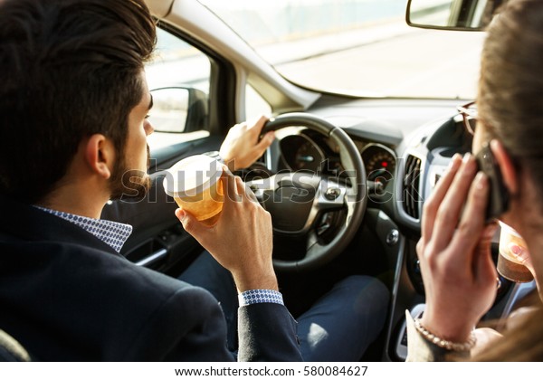 Two business friends in the car. They travel to\
business meeting.Man drives a car and drinking coffee while female\
using a phone.