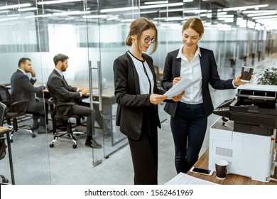 Two business ladies talking near the copier during a coffee break in the hallway of the big corporation