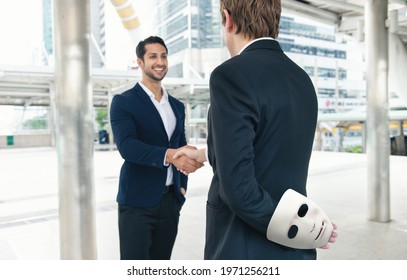 Two Business Making Handshake A Deal But Holding White Mask In His Hand Dishonest Cheating Agreement. Treachery Risk, Faking And Betray Business Partnership Concept.