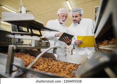 Two Business Man In Sterile Clothes Standing In Food Factory In Front Of The Production Line And Looking And Tablet. Checking The Quality Of Products And Talking.