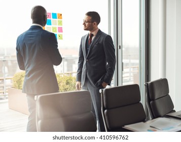 Two Business Man In Stylish Suits Standing Having A Discussion In The Office Looking At A Colorful Array Of Sticky Memos On The Window With Ideas Or A Planned Schedule