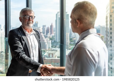 Two Business Men Discussing Their Business Together In A Lobby Of A Hotel. They Both Wearing White Shirt. The Older One Also Wear Black Jacket. The Scene Outside Is Cityscape. They Are Shaking Hand.