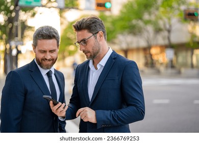 Two business men looking at messages on cellphone while walking in the street in downtown of city. Businessmen using smartphone mobile phone apps, texting message, browsing internet. - Powered by Shutterstock