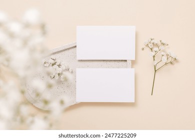 Two business paper cards with copy space on ceramic tray and beautiful gypsophila flowers on beige background, top view. Mockup paper card for design, print, greeting, wedding and invitation.