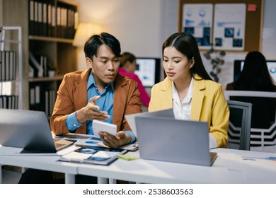 Two business partners analyzing financial data in a busy office, using a calculator and laptop, focusing on a project together - Powered by Shutterstock