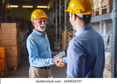 Two business partners in formal wear and with helmets on heads shaking hands in warehouse. Senior adult businessman holding tablet. - Powered by Shutterstock