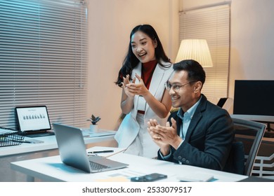 Two business people clapping their hands while looking at a laptop screen during a late night work session - Powered by Shutterstock