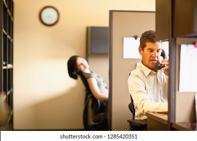 Two Business People Sit In Office Cubicles, One Talking On A Phone.
