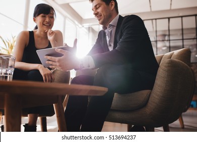 Two Business People Sitting At Lobby And Discussing Project On Digital Tablet. Young Businesswoman And Businessman Looking At Tablet Computer And Smiling.
