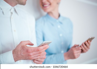 Two Business People Standing Against The Wall With Their Phones, Smiling And Looking At Each Other