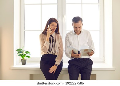 Two Business People Standing By Office Window. Woman Making A Call On Mobile Phone, Talking, Checking Information Or Organizing Event While The Man Is Reading Notes Getting Ready For Corporate Meeting