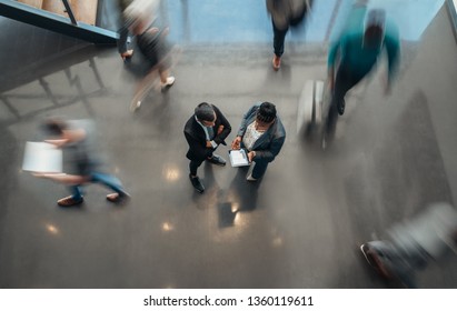 Two business people standing in the lobby of an office looking at a tablet while people are walking past in a blur