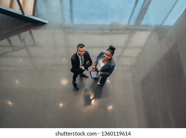 Two Business People Standing In The Lobby Of An Office Looking Up Smiling At The Camera While