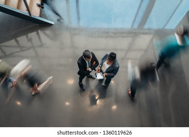 Two Business People Standing In The Lobby Of An Office Looking At A Tablet While People Are Walking Past In A Blur