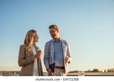 Two Business People Walking And Discussing Outside The Office. Woman Is Holding A Coffee While The Businessman Is Holding A Notebook.