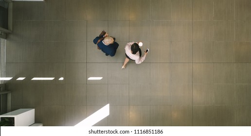 Two Business People Walking Inside Office Hallway And Talking. Top View Of Businessman And Woman In Office Building Lobby.