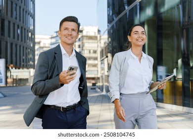 Two business professionals walk confidently outdoors, engaged with their digital devices, smiling as they move through an urban setting.

 - Powered by Shutterstock