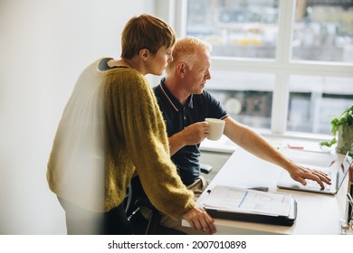 Two Business Professionals Working On Laptop Together In Office. Businessman Holding Coffee And Showing Something On Laptop To His Business Partner.