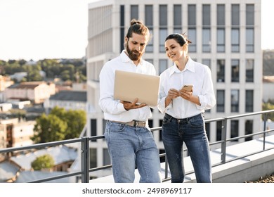 Two business professionals working on rooftop with laptop and phone. Professional attire, outdoor work setting, city skyline in background. Collaboration, teamwork, modern work and technology usage. - Powered by Shutterstock