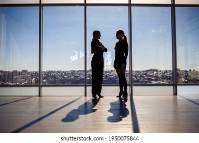 Two business women enjoying the city view and talking while standing by the large window in the office
