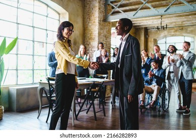 Two Business Women, One Of Japanese Ethnicity, The Other African, Shake Hands As A Multi-racial Group Of Colleagues With A Disabled Person In A Wheelchair Applaud - People And Business Concept