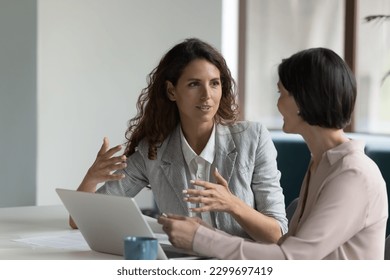 Two business women sit at desk discuss project details, diverse female colleagues met in office, share opinion, working on collaborative task, sales manager makes commercial offer to company client - Shutterstock ID 2299697419