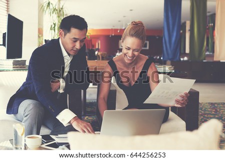 Two business workers are using laptop computer and paper documents with information about their activities in the last month before meeting with international partners, while are sitting in cafe-bar