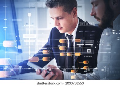 Two businessman wearing formal suit and white shirt are looking at notes, holding a smartphone and sitting in front of desktop. Office in background. Digital interface with pie diagram and globe