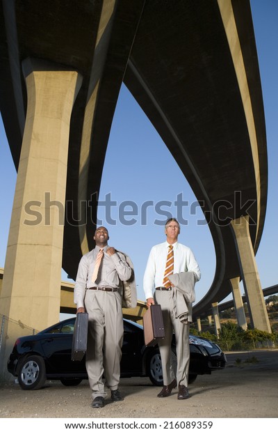 Two businessmen by car with briefcases beneath\
overpasses, low angle view