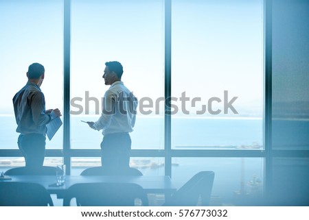 Two businessmen deep in discussion together while standing in an office boardroom with windows overlooking the ocean and city