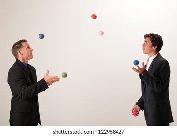 Two Businessmen Juggle Numerous Brightly Colored Balls And One Red Apple