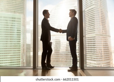 Two businessmen shaking hands standing at big window with urban cityscape, confident business partners handshaking in office, forming good relations, establish partnership, support, help. Side view - Powered by Shutterstock