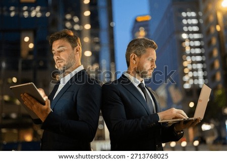 Two businessmen stand back to back analyzing data on tablet and laptop at night in New York. Male professionals reflect rivalry and determination. Scene highlights technology, business focus and urban