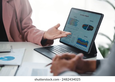Two businesspeople deeply engaged in a meeting, analyzing financial data with a tablet and paperwork adorned with various graphs and charts. Technology aids in decision-making for business growth
